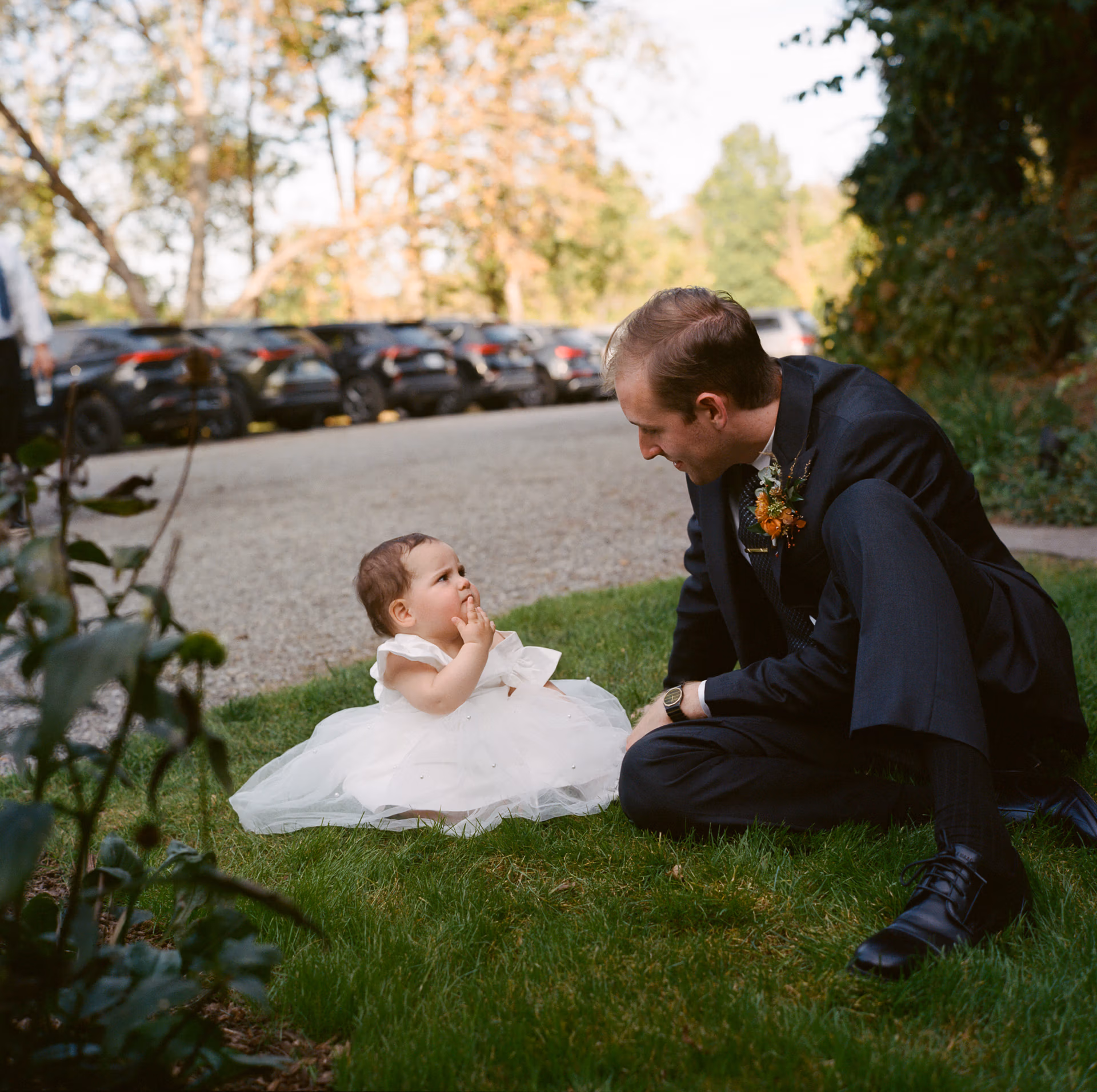 Groom in an elegant dark suit sits on a lush lawn to talk to a baby wearing a tulle dress in the grass