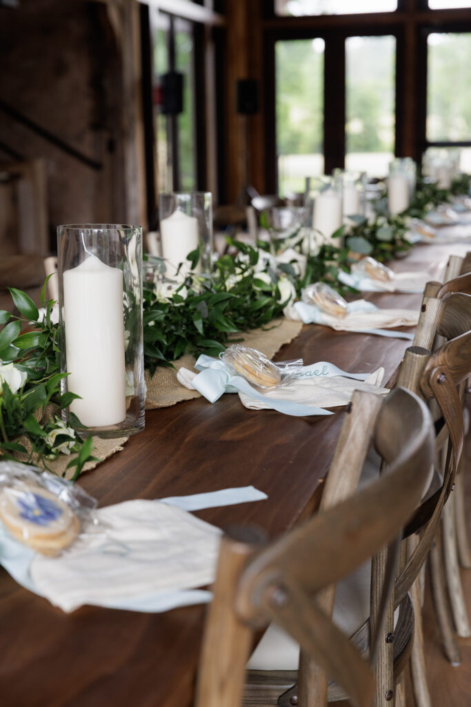 Rustic long table set with white candles in hurricanes, monogrammed white linen napkins, custom shortbread cookies decorated with pressed pansies and a table-length garland of greens