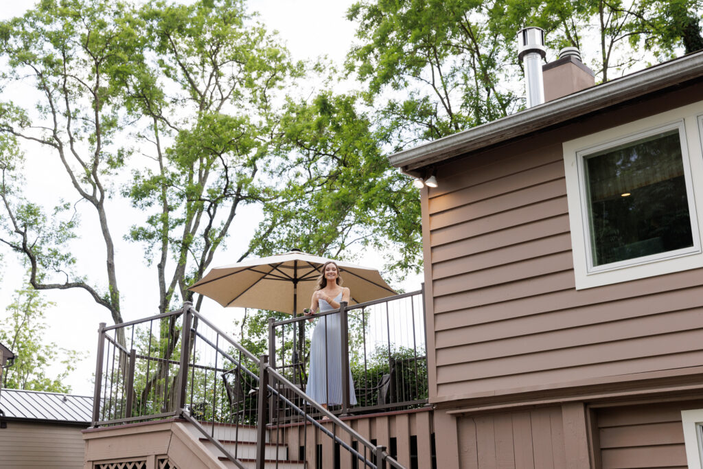 A bridesmaid in a long light blue chiffon gown smiles from the top of an elevated deck near a sun umbrella outside the bridal suite at the Woolverton Inn