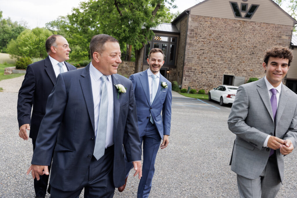 A groom in a blue suit and tie flanked by 2 brothers on his right and a guest on his left walks away from the Northridge Barn at Woolverton Inn