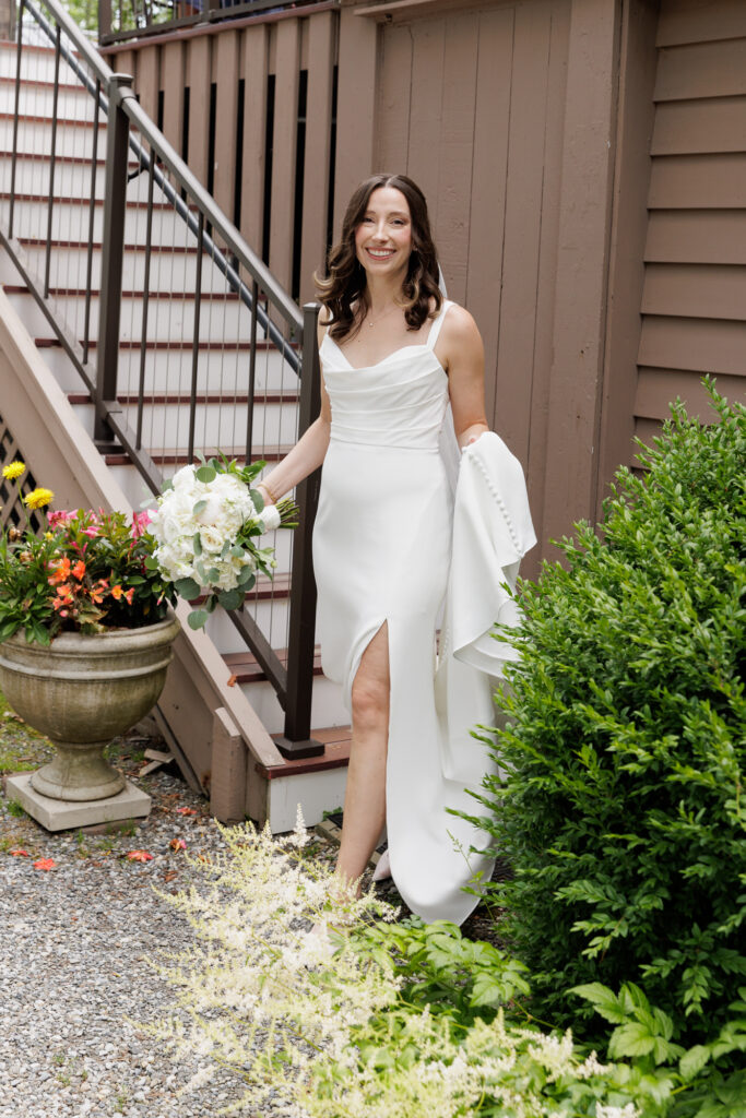 A bride with shoulder length dark hair and wearing an elegant slim satin gown holds the hem of her train in one hand and a lush white floral bouquet in the other as she descends the stairs from the bridal suite at the Woolverton Inn