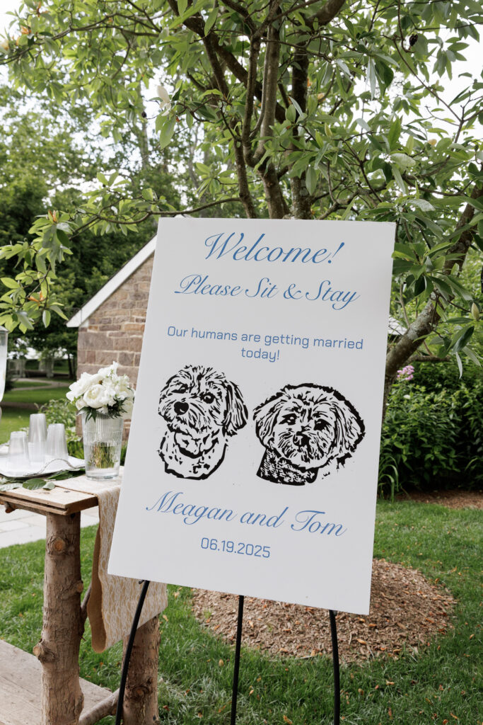 A large white sign on an easel features two dog portraits to welcome guests to a wedding 