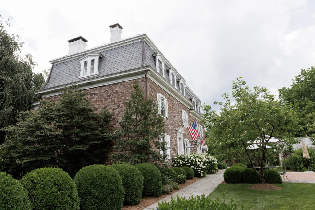 The front facade of the Woolverton Inn with lush green boxwoods lining the front walkway, and a cloudy summer sky above
