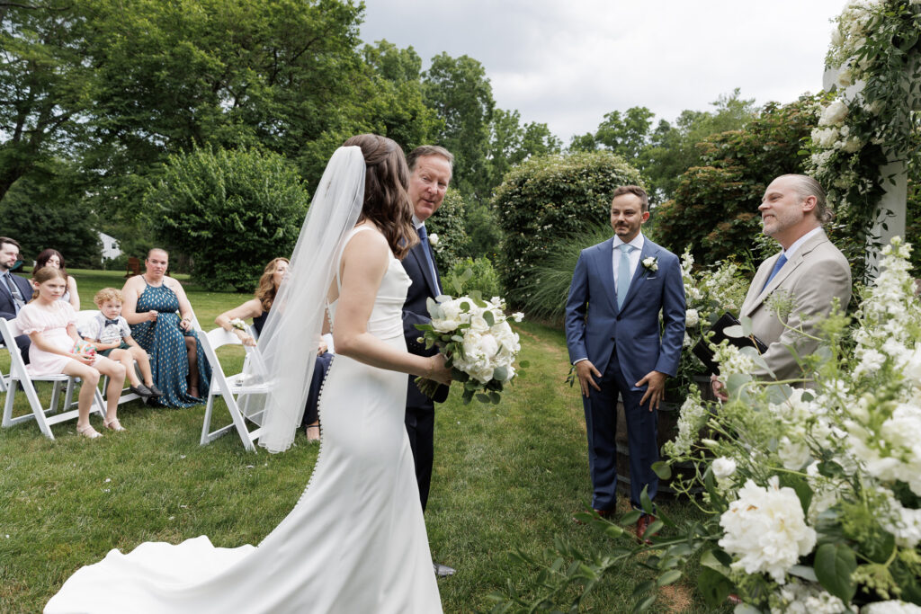A bride in an elegant slim white satin gown and veil walks arm in arm with her father during a wedding ceremony in the horseshoe garden at the Woolverton Inn