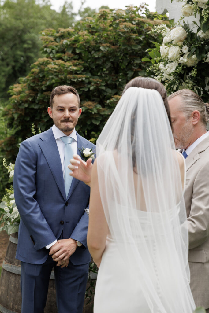 A groom in a blue suit and tie bites his lip with to prevent tears as he sees his bride at the altar
