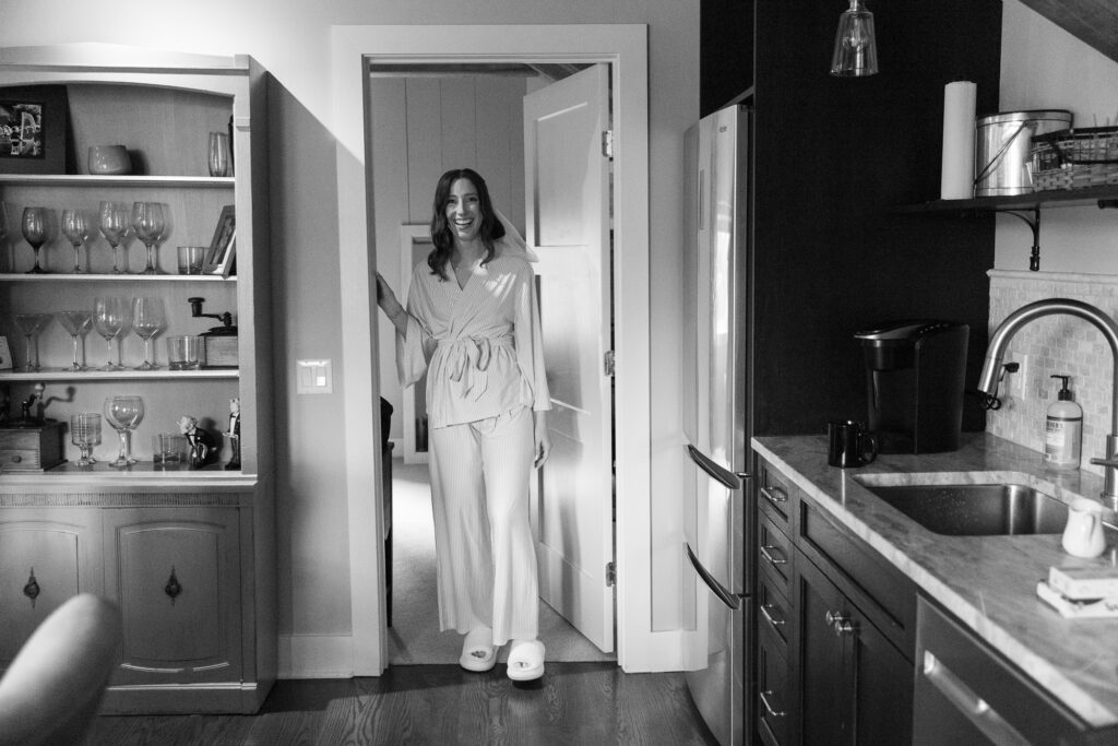 A bride with dark shoulder length hair, wearing classic button down pjs stands in a doorway in a suite at the Woolverton Inn 