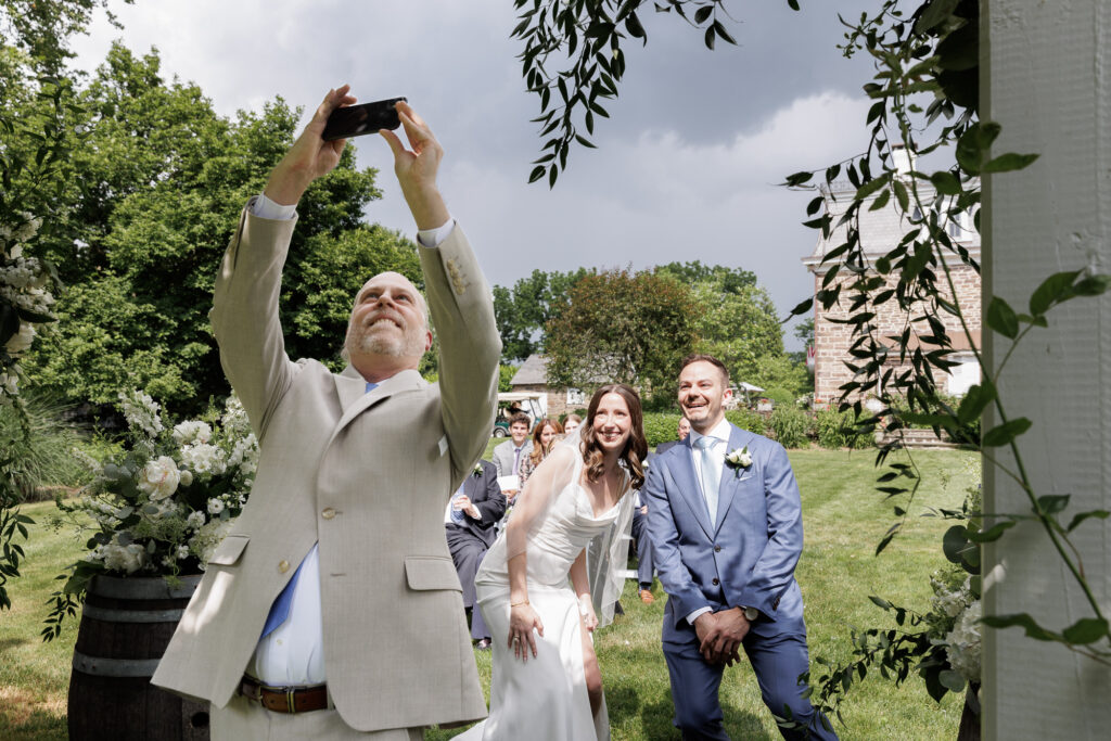 An officiant in a light tan suit turns to take a selfie to include the bride, groom and guests during a wedding ceremony at the Woolverton Inn