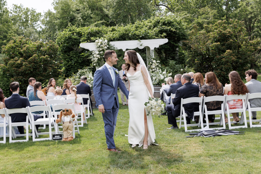 A groom in a blue suit and a bride in an elegant white slip gown turn toward each other with excitement and joy while recessing from their wedding ceremony in the horseshoe garden at the Woolverton Inn
