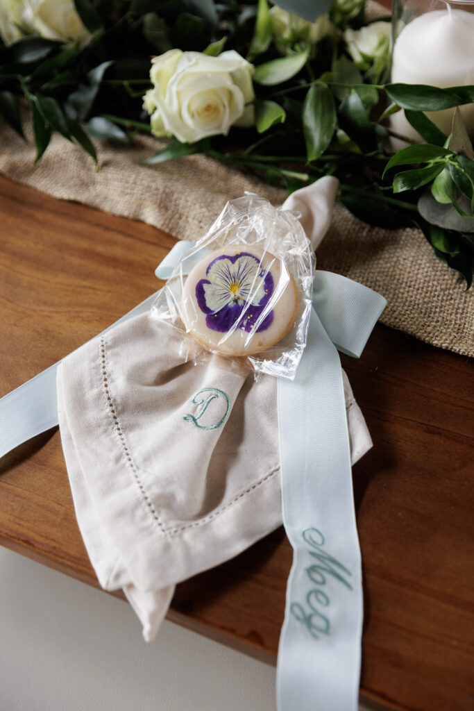 An embroidered white linen napkin tied with a customized blue satin ribbon and a shortbread cookie decorated with a pressed pansy rests at a place setting on a rustic wooden table. There is a floral arrangement of dark green leaves and ivory flowers behind it.