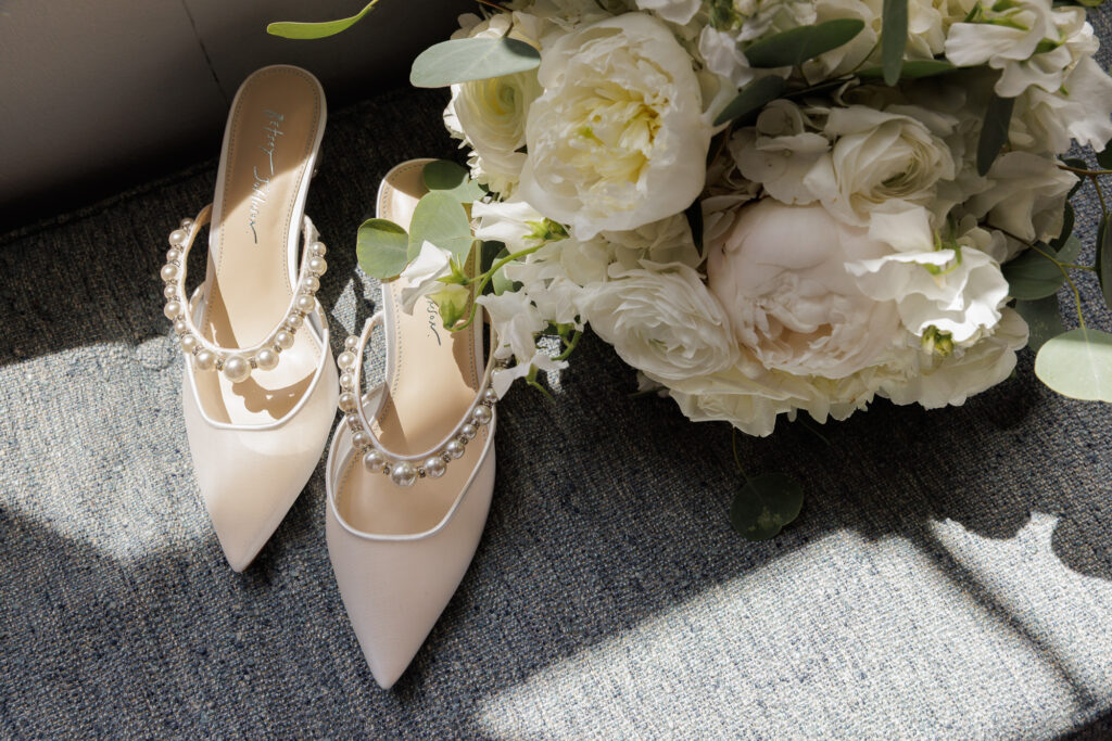 Elegant white mesh women's mules with pearl straps and a lush white bridal bouquet in dappled sun on a blue upholstered bench