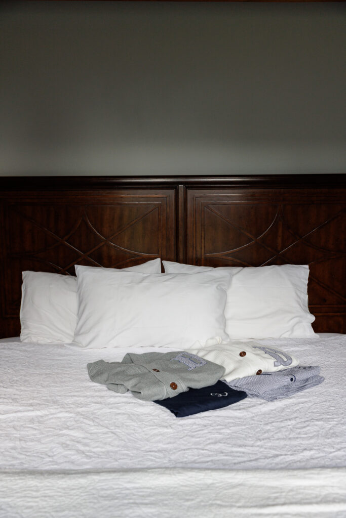 Men's gray and navy pajamas and women's white and light blue pajamas are neatly folded on an elegant dark wood bed with crisp white sheets and pillows