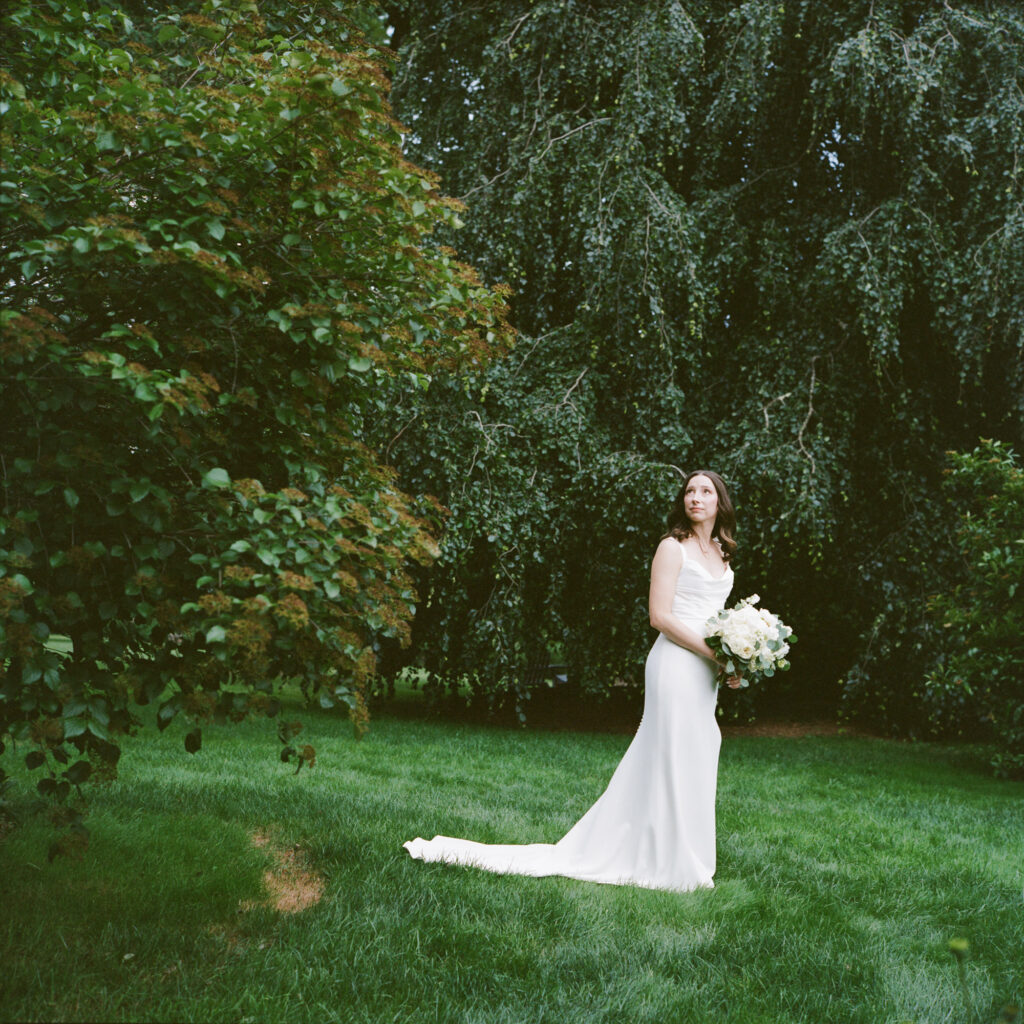 Bride with dark wavy shoulder length hair, in a long sleek modern gown holds a bouquet of elegant white flowers while standing in a lush, dark green lawn with leafy trees behind her