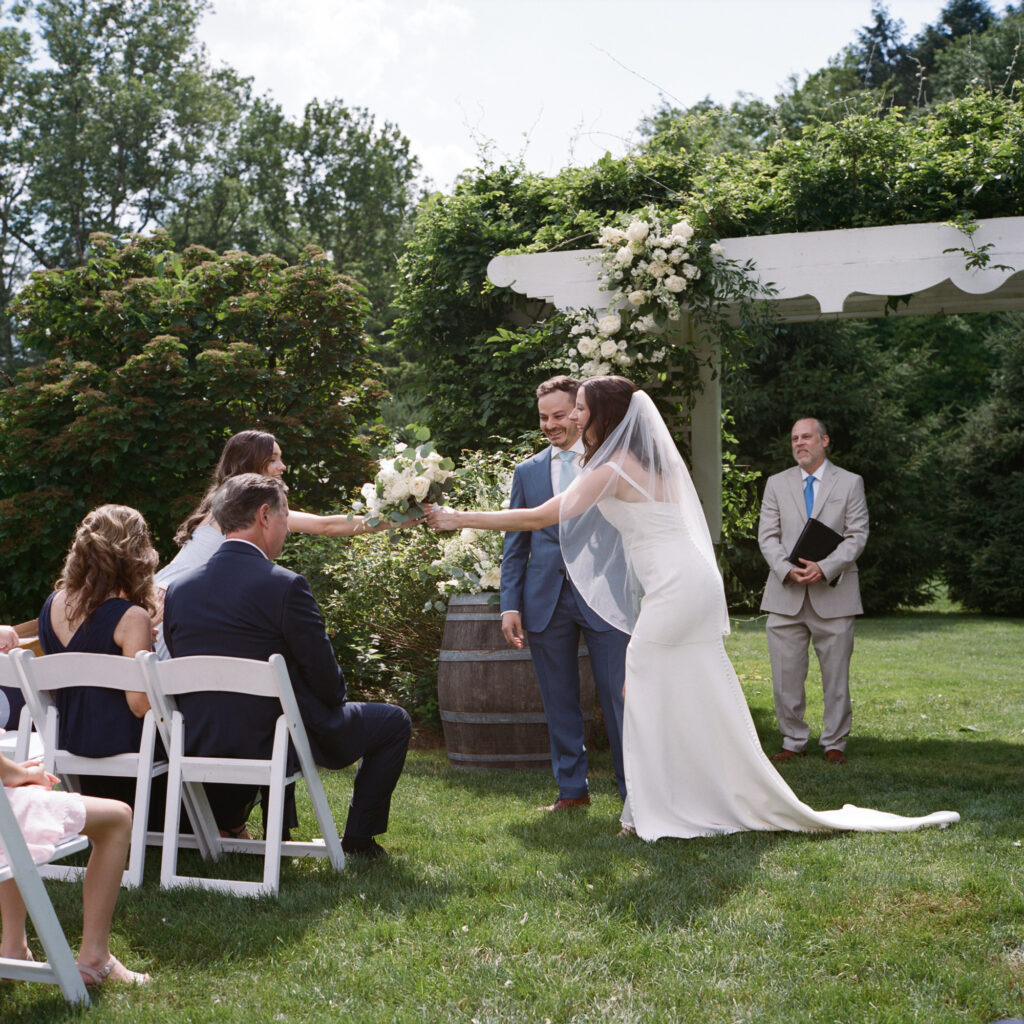 A bride stops mid recessional to grab her bouquet from the maid of honor as she and the groom exit the wedding ceremony on a sunny summer day