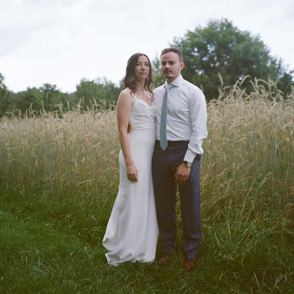 A bride in a sleek, long white gown with thin straps and a groom in a blue dress shirt, tie and dress pants embrace each other with one arm around the other's waist and pose formally without smiling while standing next to a field of tall grasses on a dark and cloudy summer evening 
