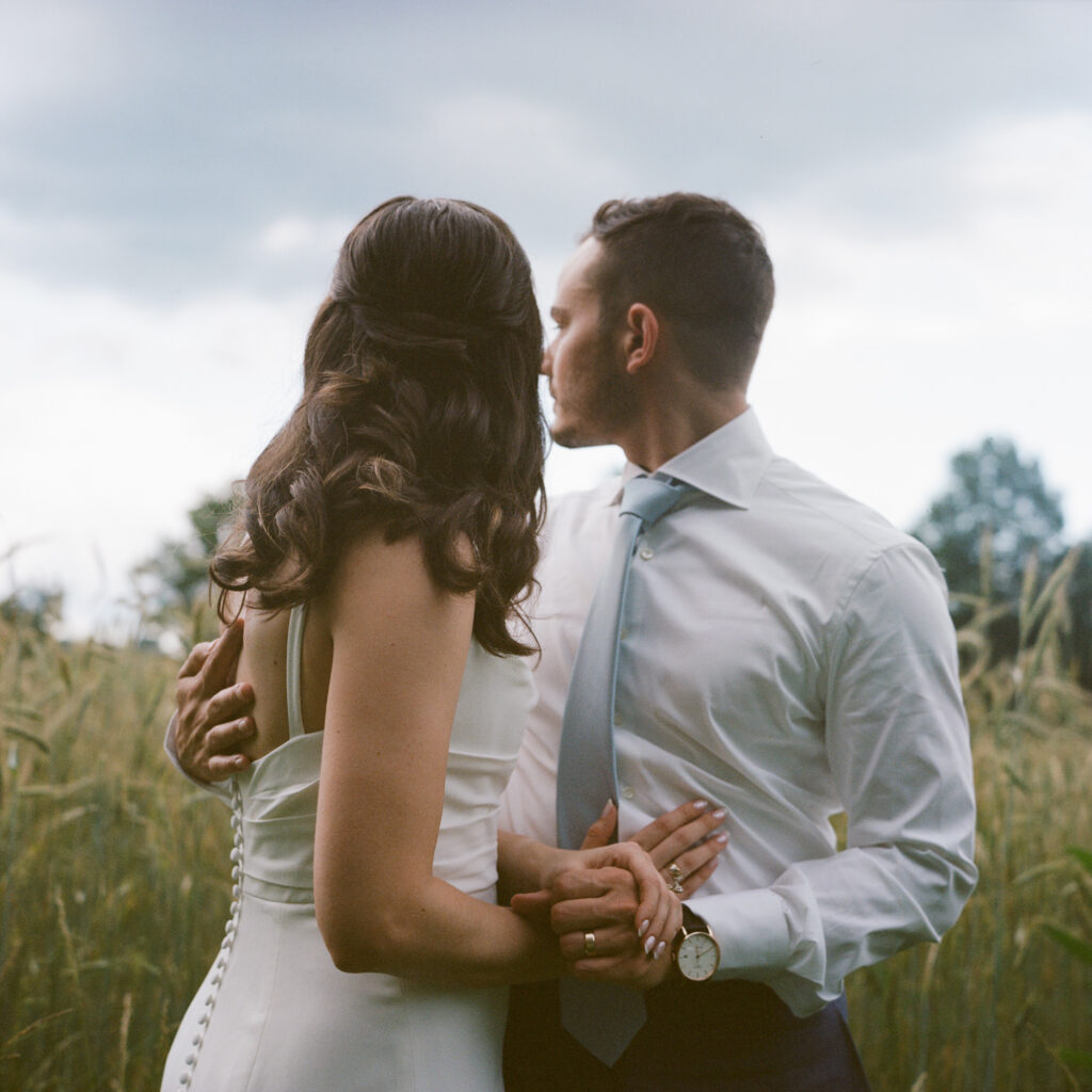 A dark cloudy summer sky looms behind a bride and groom who are embracing and looking at the horizon while standing next to a field of tall grasses