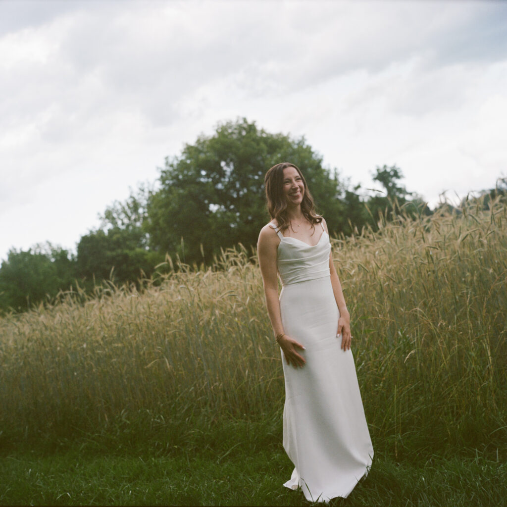 A bride in a sleek gathered satin gown stands next to a field of tall grasses while clouds gather in the summer evening sky