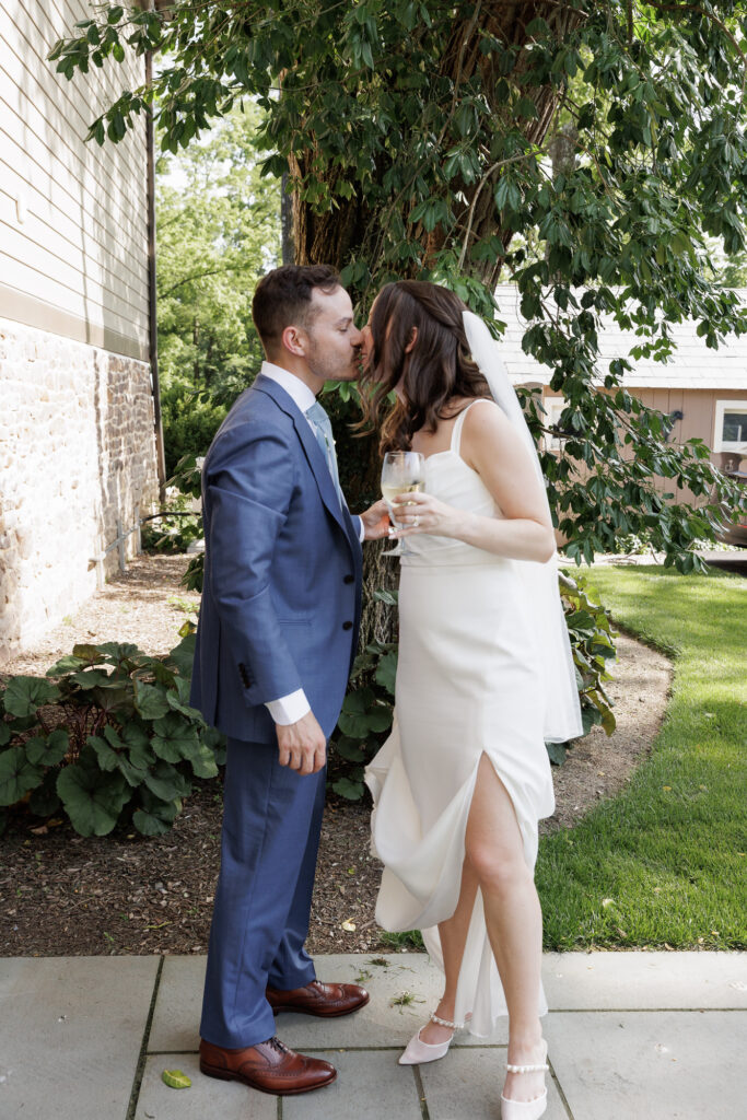 A bride casually holds up the hem of her long white gown with one hand, exposing her legs while she holds a cocktail in the other and leans in to kiss the groom, who is wearing a blue suit