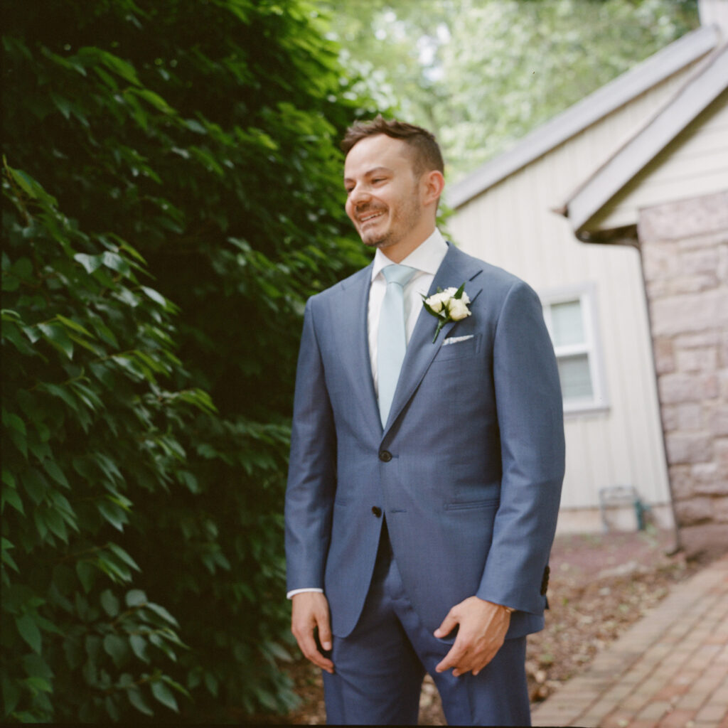 A groom wearing a blue suit and light blue tie with an ivory floral boutonniere smiles