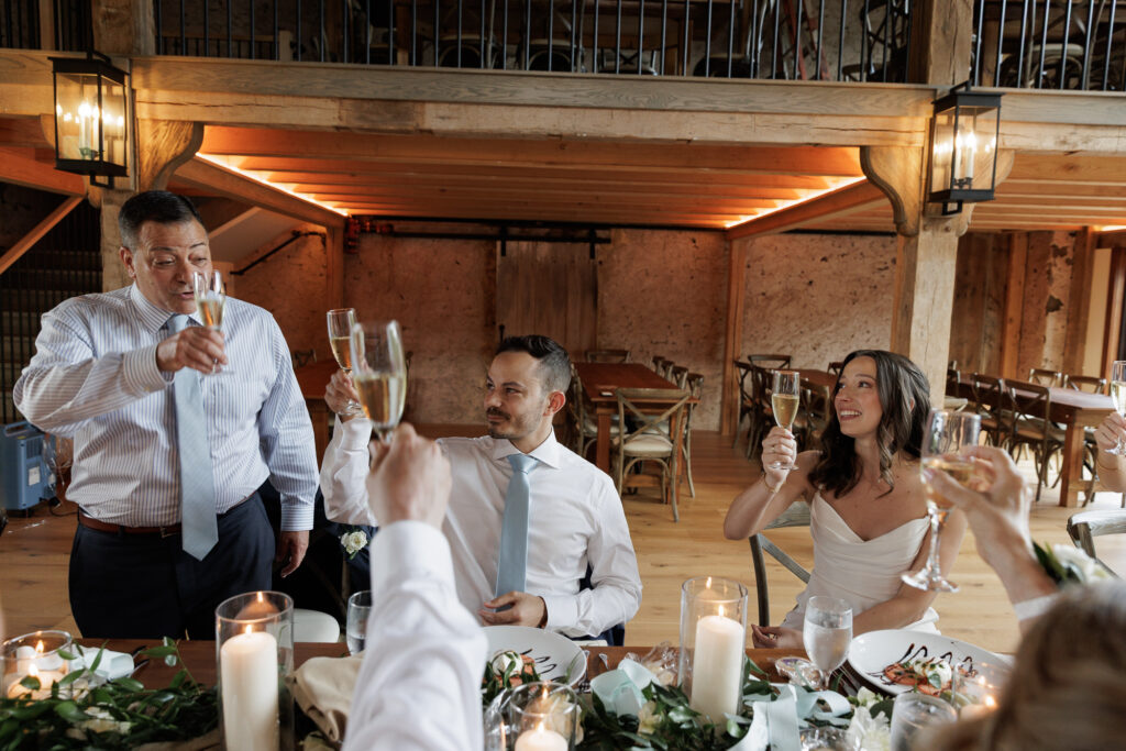 A gentleman in a shirt and tie toasts a bride and groom seated at a rustic wooden dining table inside the Northridge Barn at the Woolverton Inn