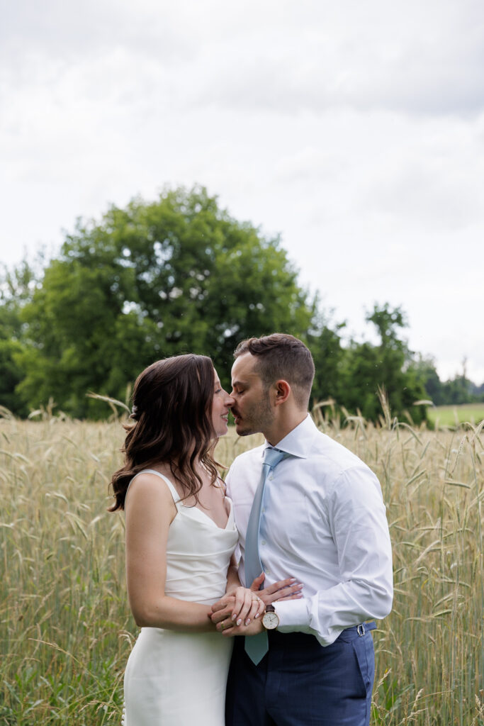 A bride in a sleeveless white gown and a groom wearing a blue dress shirt and tie kiss while standing near a field of tall grasses on a dark and cloudy summer evening