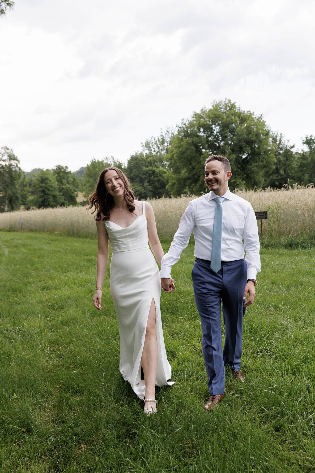 A bride in a chic, slim satin gown and groom in a blue shirt, tie and dress pants walk casually at the edge of a field of tall grasses on a cloudy summer day