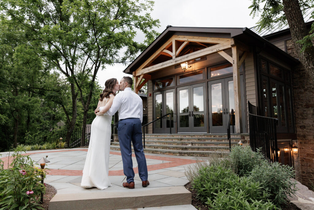 A bride and groom hold each other's hand behind the bride's back as they kiss on a flagstone patio in front of the Northridge Barn at the Woolverton Inn