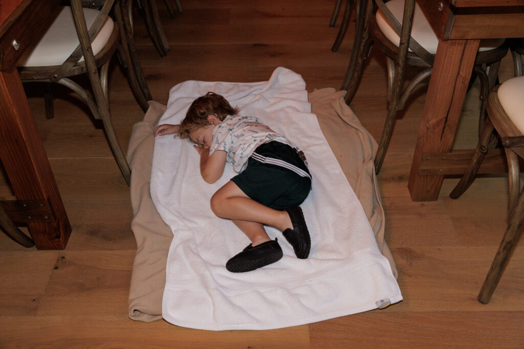 A tired little boy in a white short sleeve dress shirt, dark colored shorts and socks sleeps on top of two blankets draped on the floor between wedding reception tables 