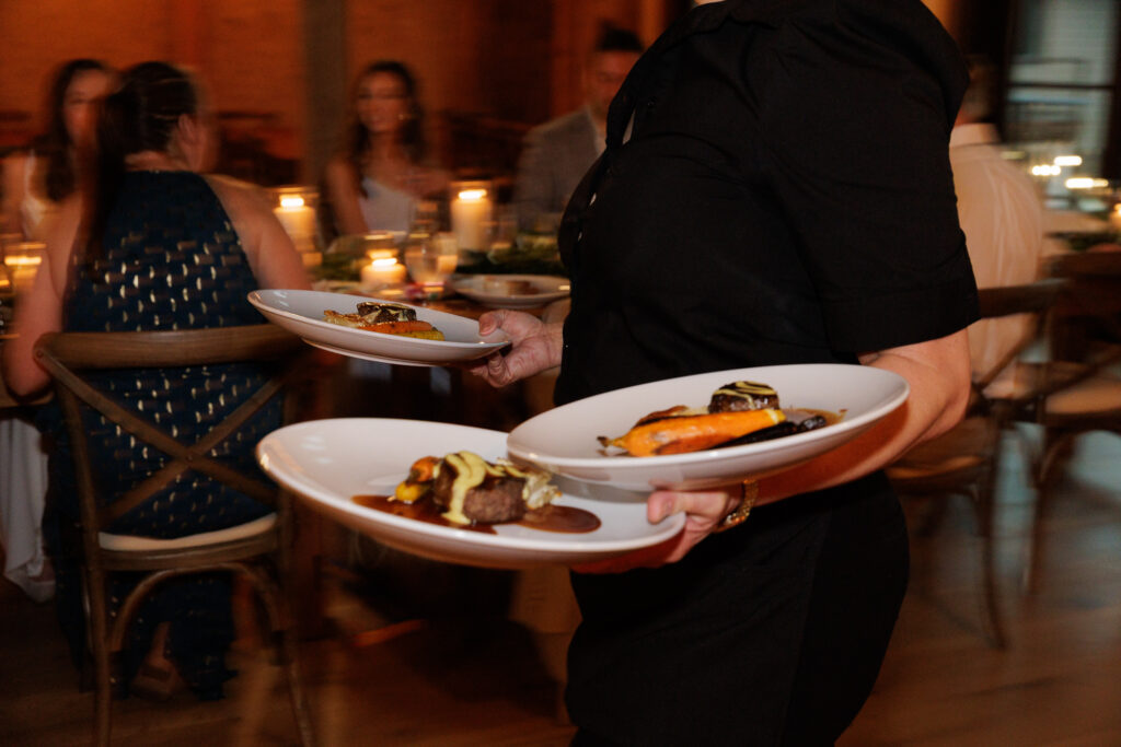 A server in black culinary attire balances two plates with dinner entrees during dinner service in the Northridge Barn