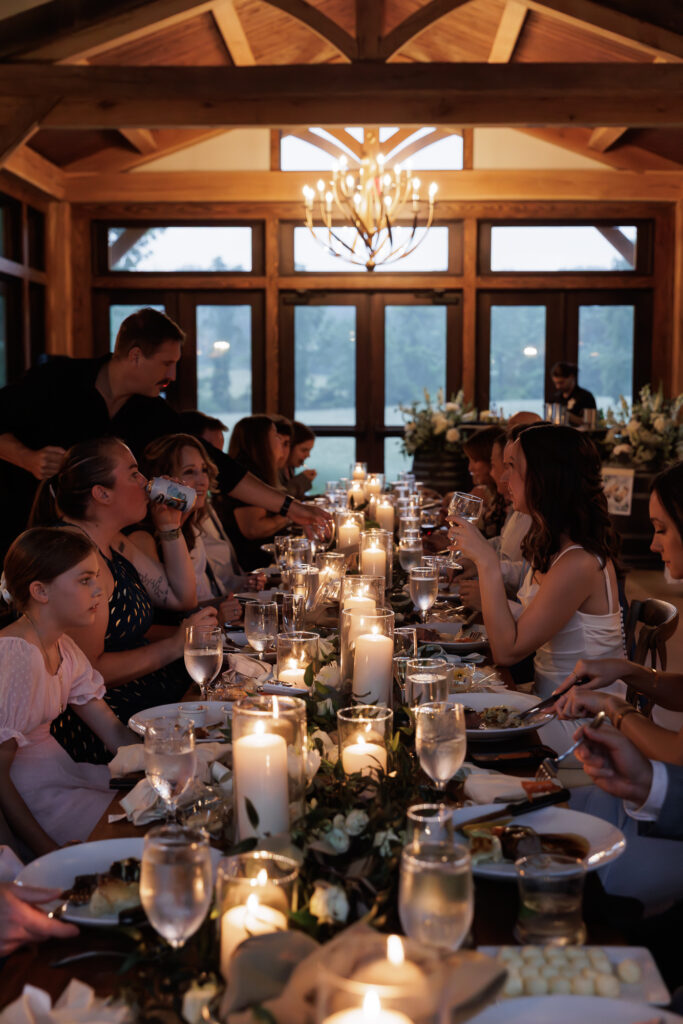 A long rustic candlelit table dressed with floral arrangements and place settings buzzes with wedding guests dining under chandeliers in the Northridge Barn