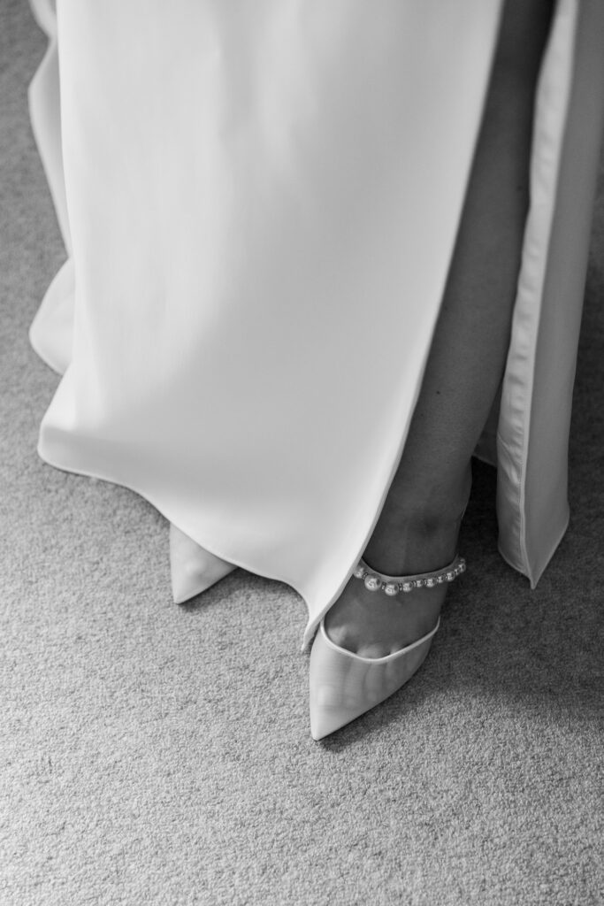 A black and white detail image of a woman wearing a slim white, floor-length gown with a high slit, a pair of pointed toe white mesh mules with a pearl strap detail peek out from under the hem