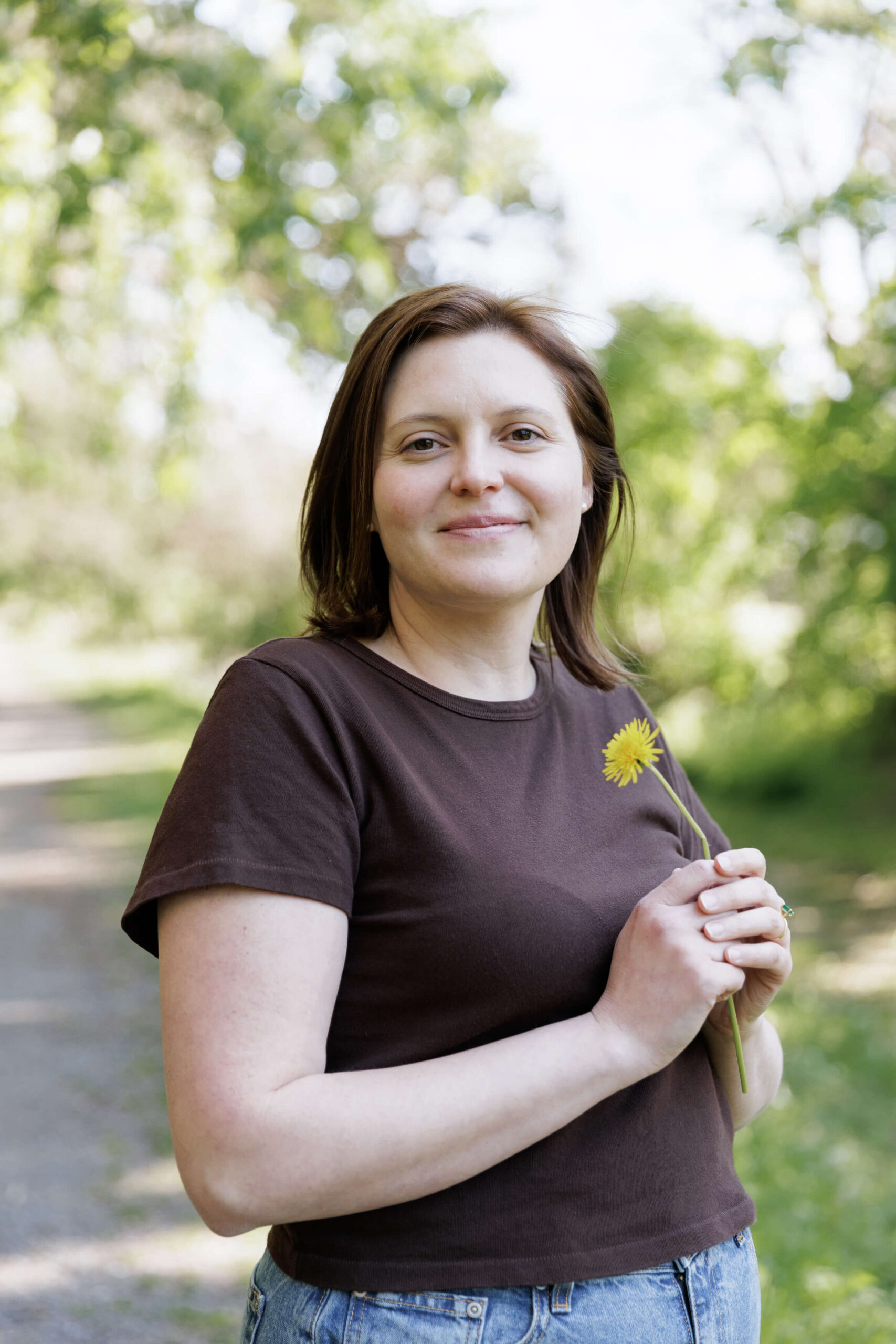 a woman wearing a dark brown t-shirt holds a single yellow dandelion in two hands and smiles