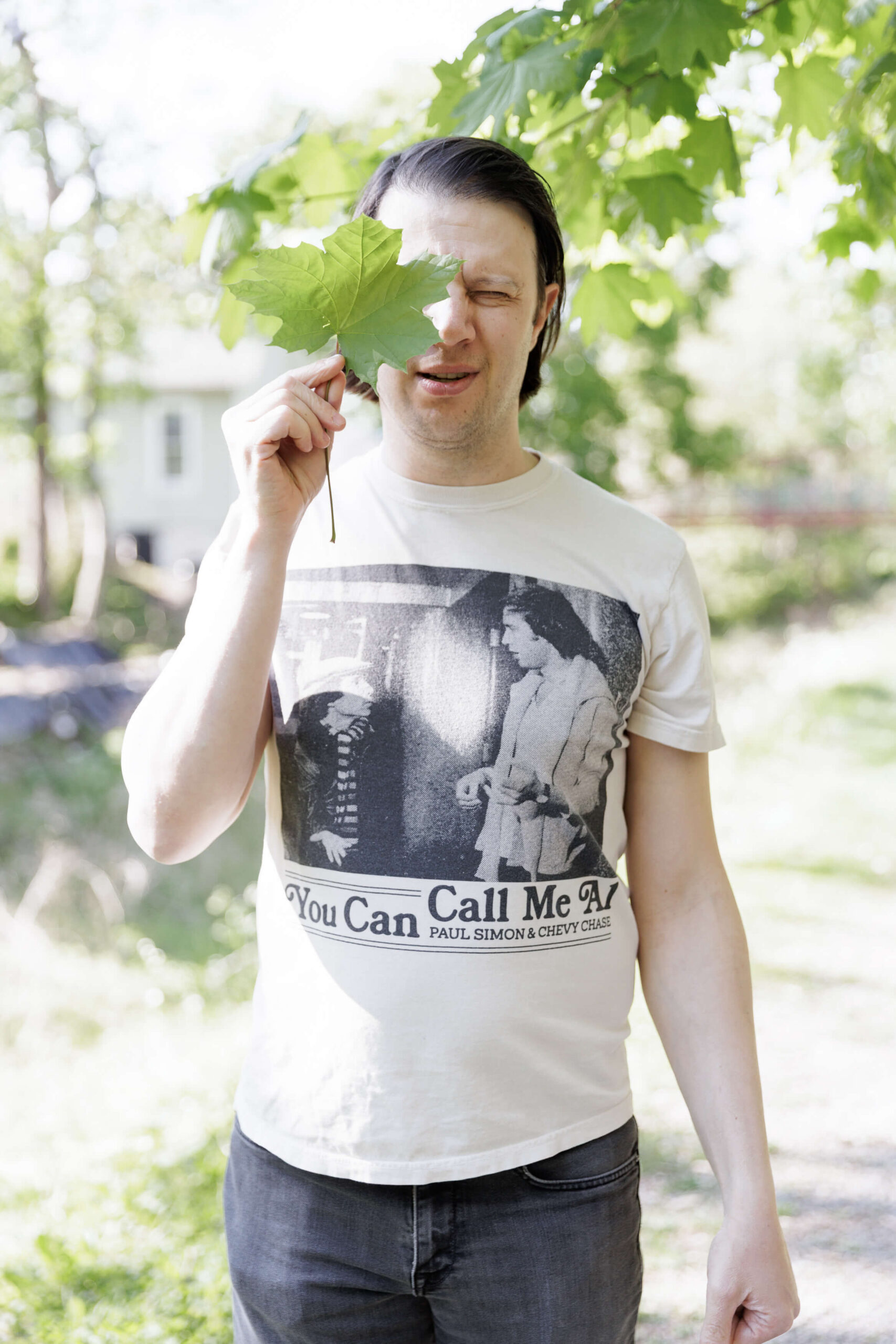 a man with dark hair wearing a vintage Paul Simon t-shirt and black jeans holds a large green maple leaf in front of his right eye, while he squints his left eye
