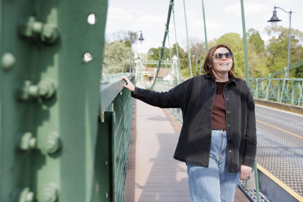 A woman with dark hair, wearing sunglasses, a button down shirt jacket over a brown t-shirt and light colored jeans holds onto the structure of the green Riegelsville suspension bridge while she smiles and looks upward on a sunny spring day