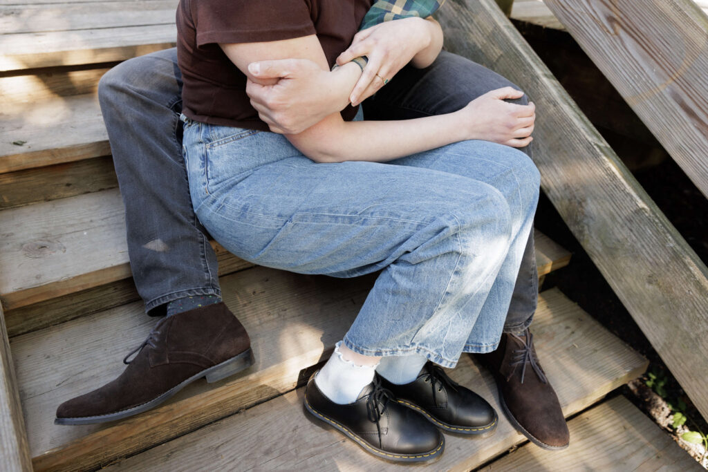 Cropped image of a man with black jeans and brown suede desert boots lovingly embracing a woman wearing a brown t-shirt and light blue jeans with black Doc Martin lace up shoes and white scalloped ankle socks as they sit on a rustic wooden stair case at a park