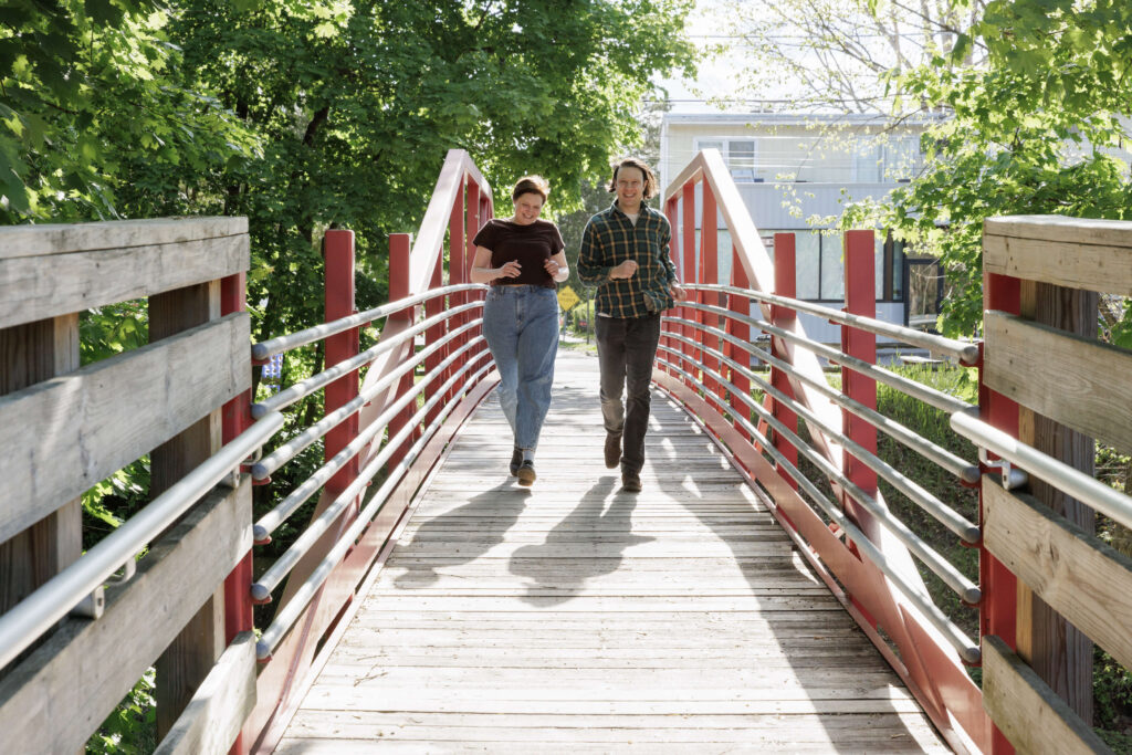A woman and a man dressed in t-shirts and jeans jog energetically across a pedestrian bridge toward the viewer