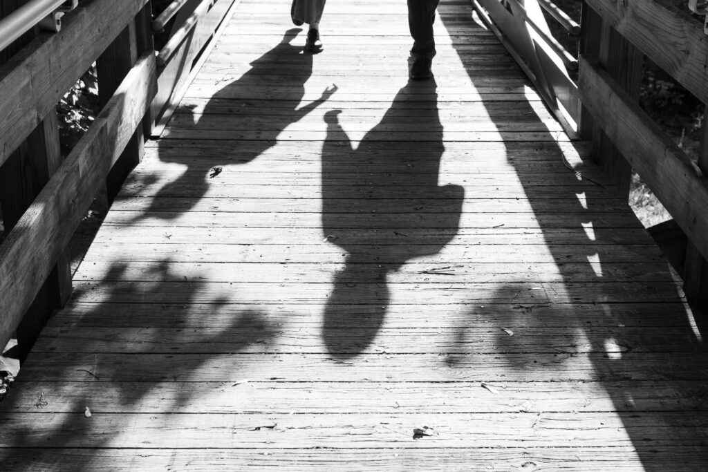black and white photo of the shadows of a couple holding hands as they walk across a wooden pedestrian bridge