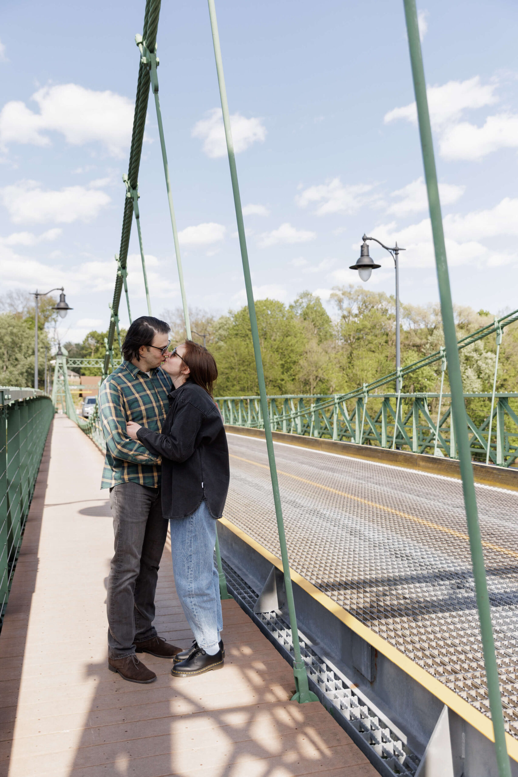A man wearing a dark green plaid shirt and black jeans kisses a woman with dark hair and sunglasses wearing a black shirt and light colored jeans. They are standing on the Riegelsville Bridge on a very sunny spring day.