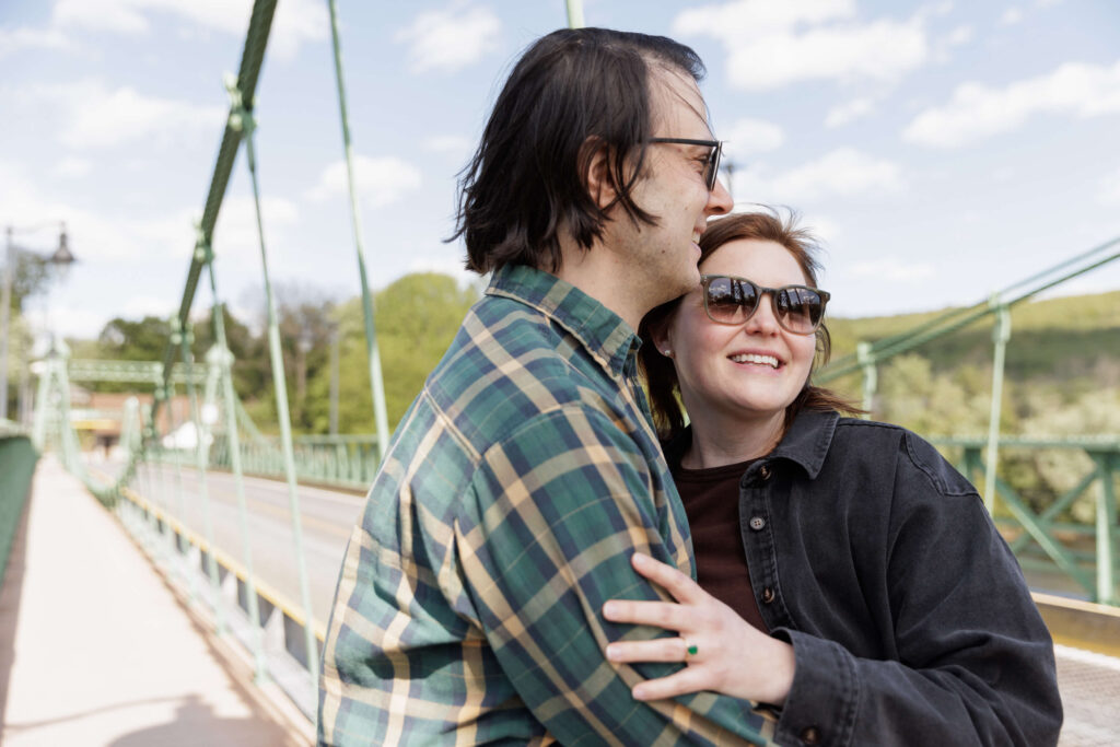 A man with dark hair wearing a green plaid shirt and sunglasses embraces a woman with dark hair and sunglasses wearing a black jacket while they stand on the Riegelsville Bridge on a bright, breezy sunny day