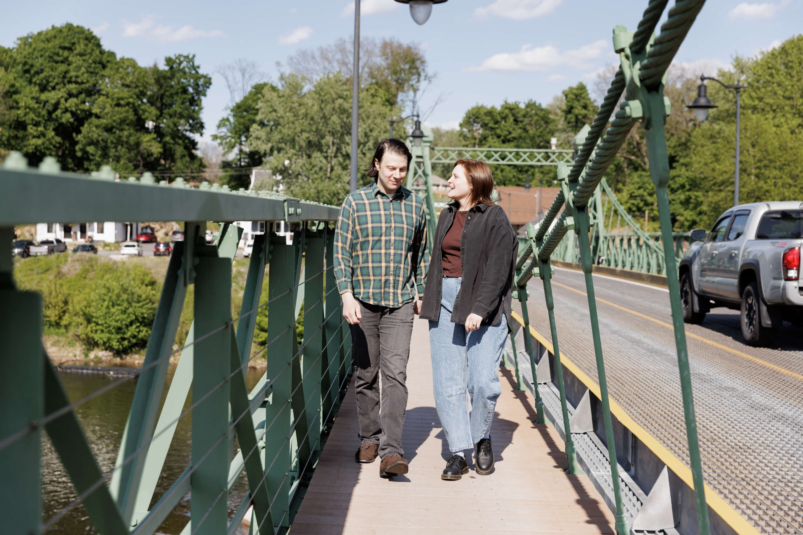 A man in a green plaid shirt and black jeans walks hand in hand with a woman with dark hair and sunglasses wearing a black jacket and light blue jeans as they cross the Riegelsville Bridge