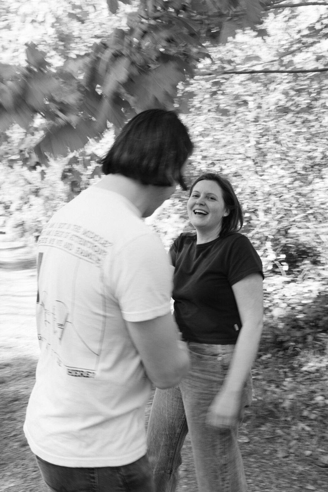 A man in a white t-shirt holds hands with a woman in a dark t-shirt and jeans has a joyous smile as they spin around in a slightly blurry black and white photo