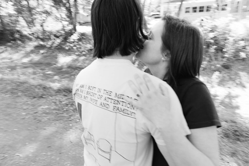 Black and white image of a man wearing a vintage Paul Simon t-shirt with the lyrics to "You Call Me Al" on the back getting a kiss on the cheek from a woman with dark hair wearing a black t-shirt