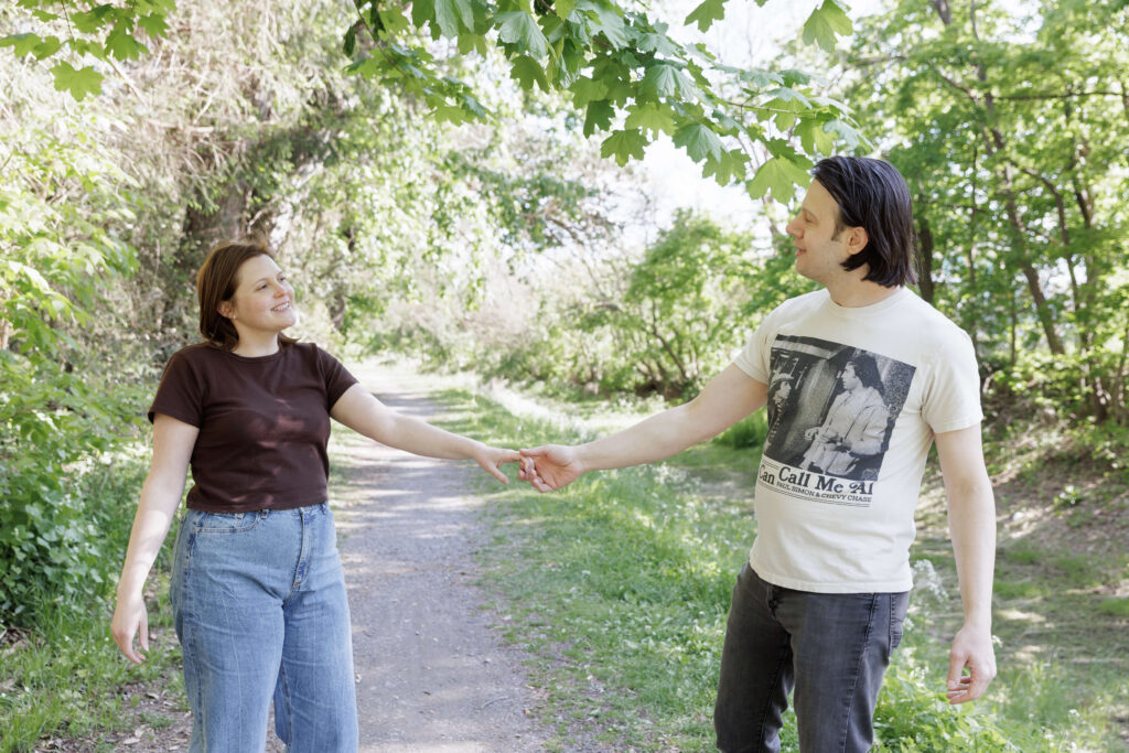 Woman wearing a dark brown t-shirt and light jeans holds hands with a man wearing a vintage Paul Simon t-shirt as they pull away and look at each other