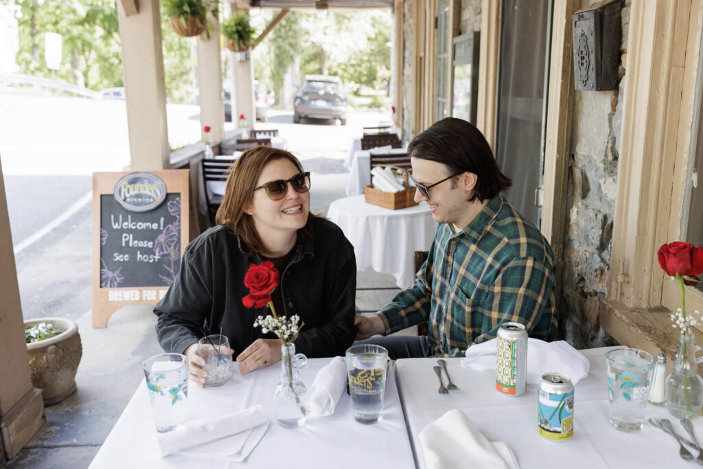 A woman in a black jean jacket wearing sunglasses and a man wearing a green plaid button down and sunglasses sit at a white tablecloth table with a red rose in a vase and chat over drinks and appetizers on the porch of the Riegelsville Inn in Bucks County PA