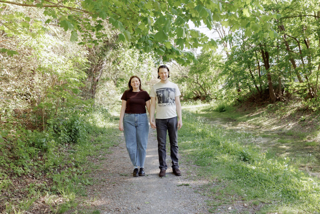 A woman in a dark brown t-shirt and light jeans stands formally next to a man wearing a vintage t-shirt and black jeans