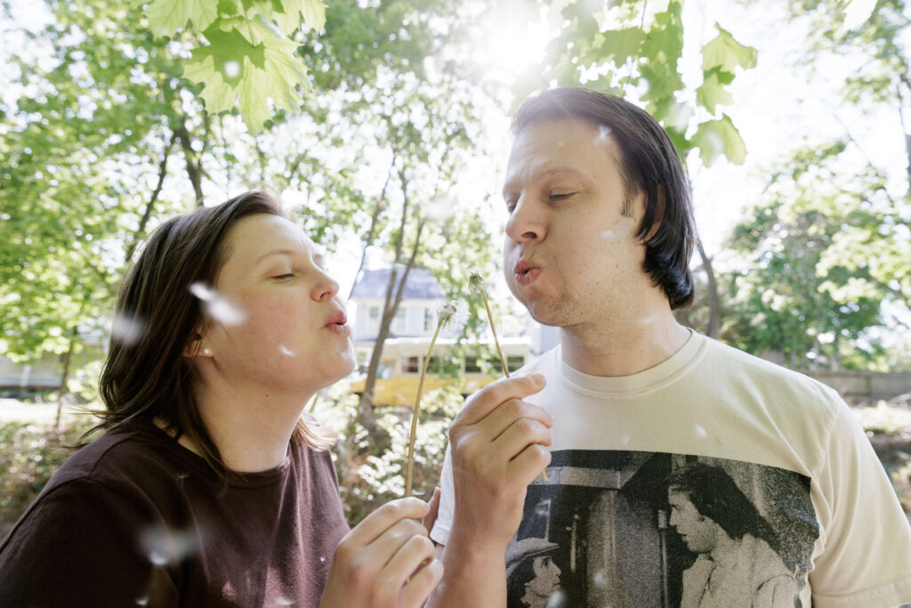 a woman in a dark brown t-shirt and a man in a vintage t-shirt playfully blow dandelion seeds towards each other while the sun shines over their shoulders on a sunny spring day