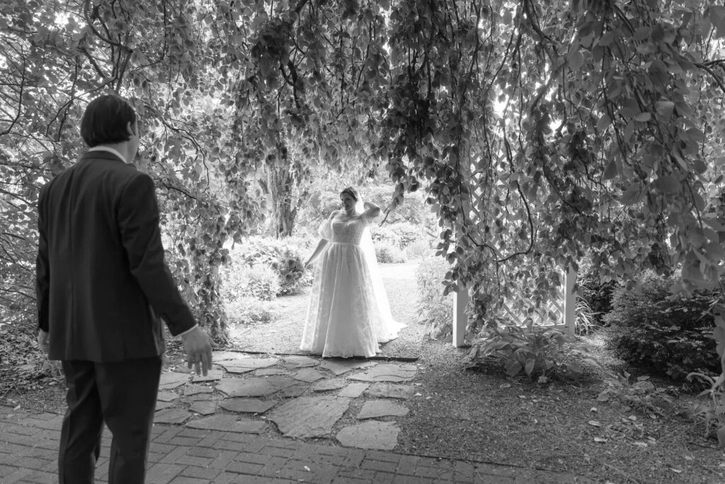 Black and white photo of a groom under a large weeping beech tree turning with joy towards a bride walking through an archway of draping branches and dappled light during a first look at the Woolverton Inn