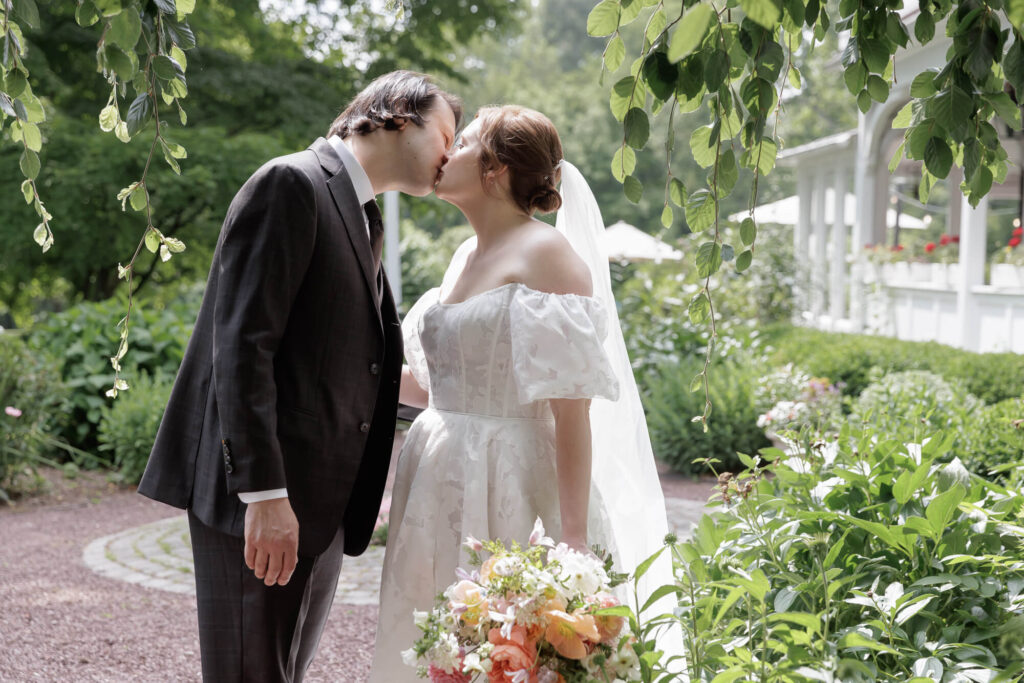 A groom kisses a bride on the cheek while they stand in a garden at the Woolverton Inn