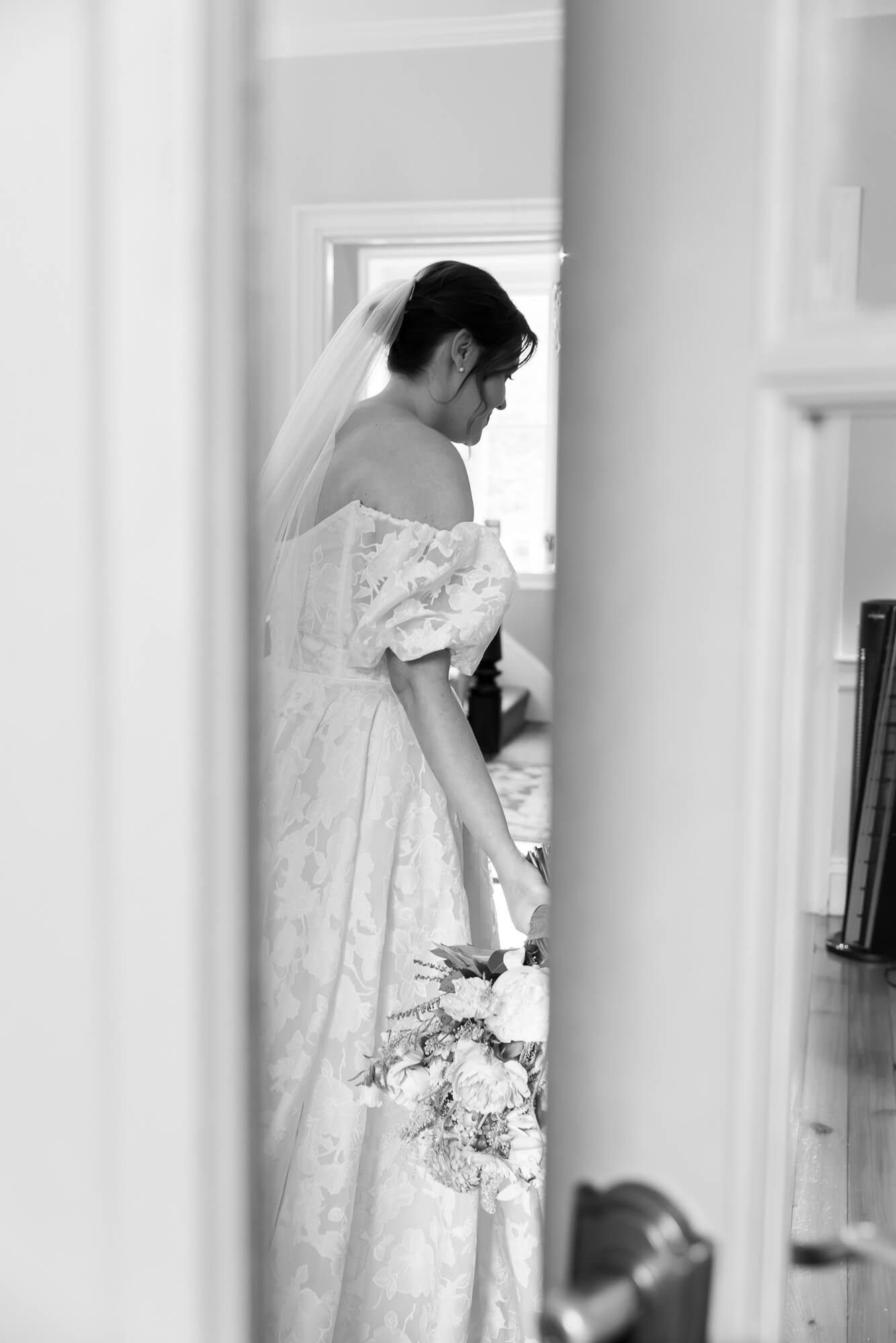 Black and white photo of a bride framed by an open door, in a quiet moment before a wedding ceremony