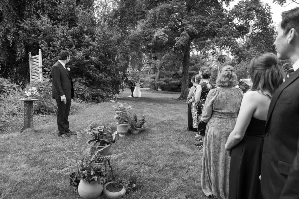 Black and white photo of the moment a bride and her dad come into view at an outdoor wedding ceremony, the groom turns to see them walking