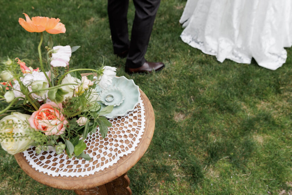 Detail shot of a small flower arrangement beside a sage green ceramic vessel holding rings on a vintage side table during an outdoor wedding in Stockton NJ